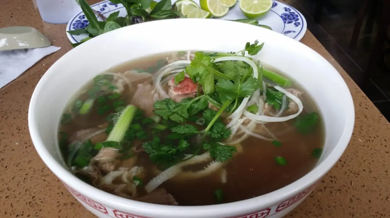 Bowl of Pho Kim Quy beef pho with rare beef, tripe, onions, and a basil-lime garnish plate